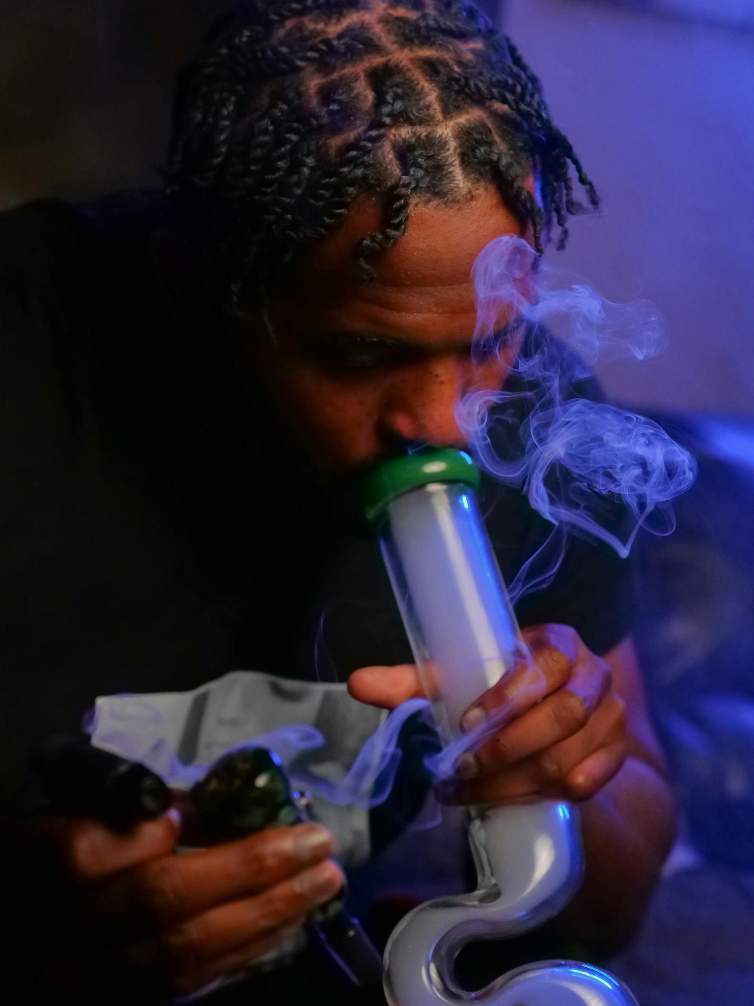 A man with dreadlocks using a bong for recreational smoking indoors.